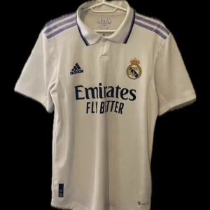 Real Madrid Vini Jr. 20 Adidas matchtröja - Säljer en officiell Real Madrid matchtröja från Adidas med Vini Jr. och nummer 20 på ryggen. Tröjan är vit med lila detaljer på axlarna, klassisk krage och broderad klubbmärke. Tillverkad i lätt och ventilerande material, perfekt för fotboll eller supporterliv. Nypris 1700