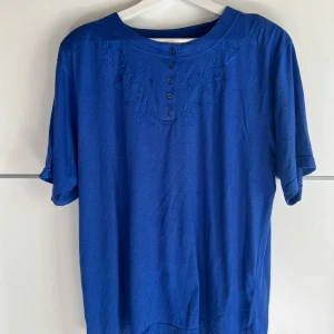 Blå t-shirt med broderi från Betty - Snygg blå t-shirt från Betty i storlek XL. T-shirten har korta ärmar, rund halsringning och dekorativa broderier framtill vid knappraden. Skön och luftig modell, perfekt för dig som gillar enkel men ändå unik stil.