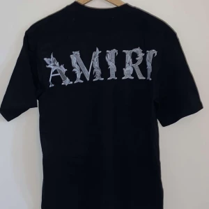 Svart AMIRI t-shirt med tryck - Svart t-shirt från AMIRI i bomull med stort, stilrent AMIRI-tryck i grått på ryggen och mindre logga på bröstet. Klassisk rund halsringning och korta ärmar. Perfekt för dig som gillar streetwear och vill sticka ut med en exklusiv vibe.