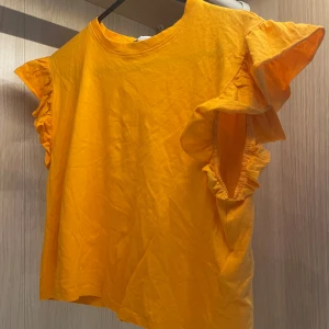 Orange volangtopp från Zara - Superfin orange topp från Zara med volangdetaljer vid ärmarna. T-shirten är i mjuk bomull och har en lös passform med korta ärmar och rund hals. Perfekt för sommaren, får dig att se brunare ut. 