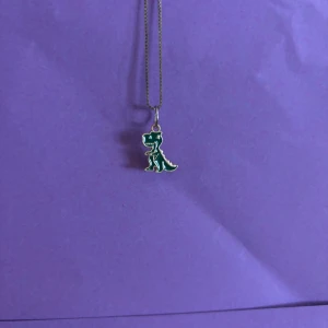 Halsband med grön dinosaurie - Sött halsband med en liten grön dinosaurie-berlock. Kedjan är tunn och silverfärgad, och berlocken har vita detaljer som framhäver dinosauriens form. Perfekt accessoar för dig som gillar lekfulla smycken med unik stil.