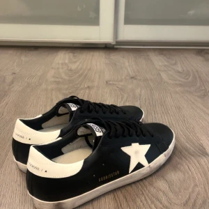Golden Goose svarta  - Golden Goose sneakers i svart skinn med vit stjärna på sidan och vit hälkappa. Klassisk låg modell med svarta skosnören och platt sula. Dom är storlek 44 och är ca 27 cm i insidan. Box medföljer ej. 