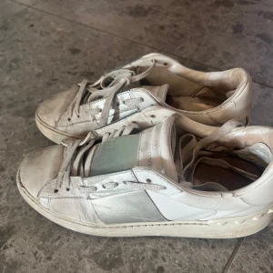 Valentino Rockstud sneakers i silver och vitt - Snygga Valentino Rockstud sneakers i vitt och silver skinn med ikoniska nitar runt hälen. Klassisk låg modell med snörning och platt sula. Perfekta för dig som gillar exklusiva detaljer och en clean look med lite edge. Passar storlek 37. Givetvis äkta och kommer med kvitto!