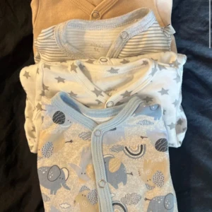 4-pack babytröjor med mönster och knappar - Fyra söta babytröjor i storlek 56. En beige, en gråvit med ränder, en vit med grå stjärnor och en ljusblå med elefanter och regnbågar. Alla har tryckknappar framtill och mjukt bomullsmaterial. Perfekt för små barn som gillar gulliga mönster.the beige one has a little tear when i have removed tag