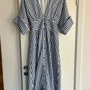 Randig långklänning med knappar - Snygg långklänning med marinblå och vita ränder, v-ringning och knappar framtill. Klänningen har trekvartsärmar med knappdetalj och ett knytband i midjan för extra passform. Perfekt för sommarens alla tillfällen. Storlek L