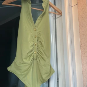 Grön baddräkt från NLY One med knappar - Snygg ljusgrön body från NLY One i storlek S. Djup v-ringning framtill med dekorativa knappar och rynkade detaljer längs magen. Öppen rygg och i stretchigt material. 