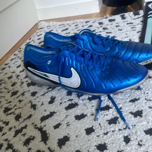 Nike Tiempo blå fotbollsskor - Snygga blå Nike Tiempo fotbollsskor med silverfärgade detaljer och stor vit swoosh på sidan. Skorna har snörning, rund tå och platt sula med dobbar för bra grepp på planen. Ovandelen är i syntet och mesh för bra andningsförmåga och passform.