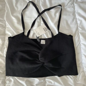 Svart croppad linnetopp med twist - Svart croppad linnetopp från H&M med smala axelband som korsas i ryggen. Toppen har en snygg twist-detalj framtill som ger en cool look. Perfekt till höga jeans eller kjol. Materialet känns mjukt och stretchigt