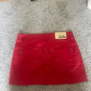 Lågmidjad jeanskjol  - Snygg röd kortkjol från Bisou D'ine med klassisk jeanslook, fem fickor och bälteshällor. Kjolen har knapp och dragkedja framtill samt coola nitar vid fickorna. Perfekt för dig som vill sticka ut med färg!