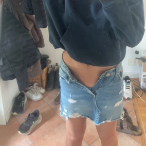 Blå jeanskjol med slitningar - Säljer en blå jeanskjol med råa kanter och slitna detaljer. Kjolen är kort och har klassisk femficksdesign med knapp och dragkedja framtill. Perfekt för dig som gillar en avslappnad och trendig stil. Är en XS men sitter mer som en XXS