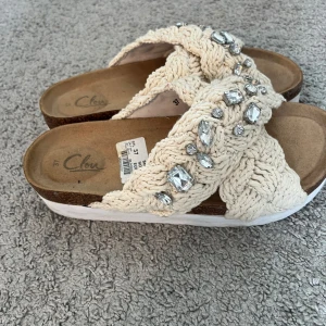 Beige sandaler med stickade band och stenar - Snygga sandaler från Clou i beige med breda, stickade band i kors och glittrande stenar. Nypris 450kr,  Skorna har en platt sula och öppen tå, perfekt för varma dagar✨Använda några fåtal gånger.