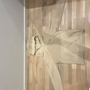 Beige hoodie från GANT med brodyr - Snygg beige hoodie från GANT med broderad logga på bröstet. Klassisk känguruficka framtill och huva. Perfekt för chill dagar och enkel att matcha med jeans eller joggers.