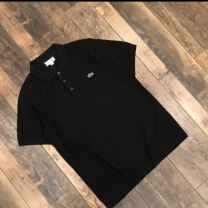 Svart pikétröja från Lacoste - Klassisk svart pikétröja från Lacoste med korta ärmar och ikoniska krokodilloggan på bröstet. Den är ej använd och kommer i storlekar s,m,l,xl 