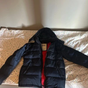 Mörkblå dunjacka från Tommy Hilfiger - Snygg mörkblå dunjacka från Tommy Hilfiger med huva och röd insida. Jackan har quiltad design, dragkedja framtill och långa ärmar. Perfekt för kalla dagar när du vill hålla stilen och värmen.