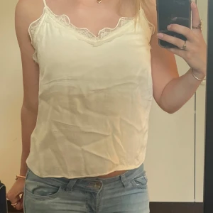 Vit linnetopp med spetsdetaljer - Säljer en söt vit linnetopp med tunna axelband och v-ringning. Toppen har fina spetsdetaljer längs ringningen och är perfekt att styla med jeans eller kjol för en fräsch look!💞💞