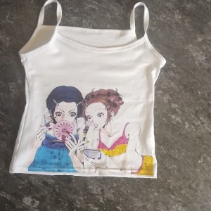 Vit linne-topp med anime-tryck - Vit ärmlös linne-topp med smala axelband och ett stort färgglatt anime-inspirerat tryck framtill som visar två tjejer i färgstarka kläder. Toppen har en rak passform och är gjord i mjukt material, perfekt för en cool och avslappnad stil.