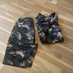Kläder  - Säljer ett camo-set från Warp med kortärmad skjorta och matchande cargoshorts. Setet har grön, beige och svart camouflage-mönster och är gjort i bomull. Skjortan har klassisk krage och knappar, shortsen har dragkedja och flera fickor. Perfekt för dig som gillar streetstyle.