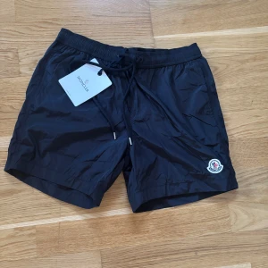 Svarta badshorts från Moncler - Svarta badshorts från Moncler med elastisk midja och snörning. Klassisk Moncler-logga framtill och en bakficka med dragkedja och rödvit detalj. Lätta och snabbtorkande, perfekta för stranden eller poolen.
