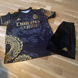 Real Madrid Adidas fotbollströja svart/guld - Snygg Real Madrid fotbollströja från Adidas i svart med guldiga detaljer och drakmönster. Kortärmad modell med klubbmärke och sponsortryck framtill. Tillverkad i lätt och ventilerande material, perfekt för träning eller match. Det finns olika stårlek och pris. Fråga om pris 