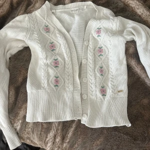 Vit kofta med broderade blommor Holly & Whyte - Supersöt vit kofta från Holly & Whyte med broderade rosa blommor och gröna blad framtill. Koftan har kabelstickade detaljer, ribbade muddar och knappar hela vägen.