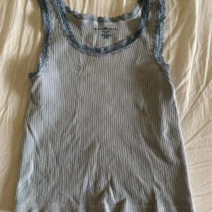 Randig ribbad linnetopp med spets - Supersöt ljusgrå och vit randig linnetopp från Brandy Melville. Toppen är ribbad och har fina spetsdetaljer i grått längs urringning och ärmhål. Perfekt att styla med jeans eller kjol för en chill look.