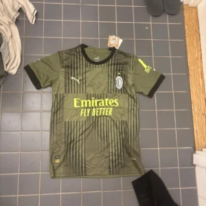 AC Milan Ibrahimović tröja Puma S - AC Milan fotbollströja från Puma i olivgrönt med svarta vertikala ränder och svarta detaljer vid krage och ärmslut. Tryck med Ibrahimović 11 på ryggen och klubbmärke på bröstet. Tillverkad i lätt och ventilerande polyester, perfekt för match eller träning.