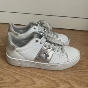 Vita sneakers från U.S. Polo Assn. - Snygga vita sneakers med silvriga detaljer och U.S. Polo Assn.-logga på sidan. Skorna har snörning, platt sula och rund tå. Perfekta för en sportig och stilren look. Tillverkade i syntetmaterial med inslag av skinnimitation.