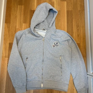 Grå Moncler hoodie med dragkedja - Snygg ljusgrå hoodie från Moncler med dragkedja framtill och två fickor med dragkedja. Tröjan har huva och ett broderat Moncler-märke på bröstet. Tillverkad i mjuk bomullsmix som är perfekt för chill dagar. Passar dig som gillar stilren och sportig look.