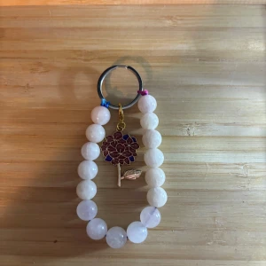 Nyckelring med blomberlock - Snyggt armband med stora vita pärlor och en charmig blomberlock i lila och guld. Perfekt accessoar för att ge din stil en söt och färgglad touch. Armbandet har elastisk tråd och detaljer i metall.