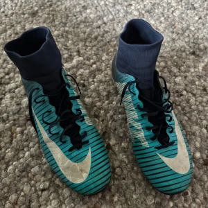 Nike Mercurial blå fotbollsskor - Snygga Nike Mercurial fotbollsskor i blått med svarta och vita ränder, stor vit swoosh och högt skaft i stretchigt tyg. Skorna har snörning och platt sula med dobbar för gräsplan. Perfekta för dig som vill sticka ut på planen. Skorna är bra skick men inte tvättade! Storlek 45