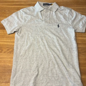 Grå pikétröja Polo Ralph Lauren XS - Snygg ljusgrå pikétröja från Polo Ralph Lauren i modellen Custom Slim Fit. Klassisk krage, korta ärmar och broderad marinblå logga på bröstet. Perfekt basic till garderoben och enkel att matcha med allt. Tillverkad i mjuk bomull för skön känsla.