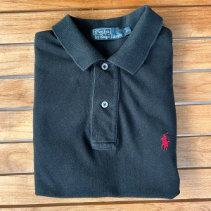 Ralph Lauren Pikè Svart S - Klassisk svart pikétröja från Ralph Lauren med röd broderad logga på bröstet. Tröjan har korta ärmar, krage och knappar framtill. Tillverkad i 100% bomull för en skön och luftig känsla. Perfekt för en stilren och avslappnad look.