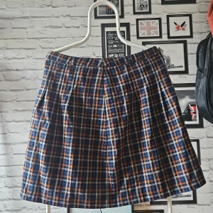 Rutig kortkjol i blått och orange - Snygg kortkjol med rutigt mönster i blått, orange, svart och vitt. Kjolen har dragkedja bak och är gjord i ett mjukt polyestertyg. Perfekt för dig som gillar en preppy och färgstark stil.