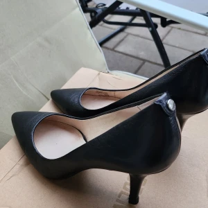 Svarta pumps från Michael Kors - Stilrena svarta pumps från Michael Kors i skinn med spetsig tå och smal klack. Snygg detalj med silverfärgad logoknapp bak på hälen och metallicfärgad innersula. Perfekta för dig som gillar klassisk och elegant stil.