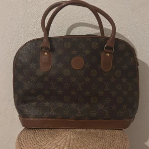  brun monogram handväska - Snygg handväska  i klassiskt monogrammönster. Väskan är brun med ljusbruna detaljer och handtag i skinn. Den har rundad form och dragkedja upptill. Perfekt för dig som gillar lyxiga accessoarer med ikonisk design. Se defekterna på bilderna💕