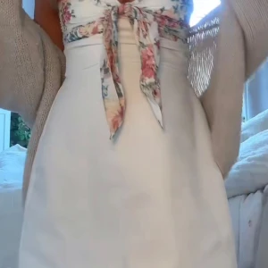 Vit kortklänning med blommigt knyt - Supersöt vit kortklänning med blommigt mönster upptill och knyt framtill. Klänningen är i storlek xs