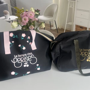Blommig shoppingväska Le Temps des Cerises - Stor svart shoppingväska från Le Temps des Cerises med ljusrosa handtag och dragkedja. Väskan har ett gulligt blommönster i rosa och grönt samt tryckt logga framtill. Perfekt för att bära med sig allt man behöver till skolan eller stan.