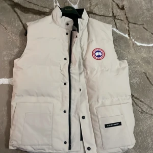 Beige dunväst från Canada Goose - Snygg beige dunväst från Canada Goose med klassisk logga på bröstet. Västen har hög krage, två stora fickor framtill och stängs med både dragkedja och knappar. Perfekt för lager-på-lager och håller dig varm under kyliga dagar.