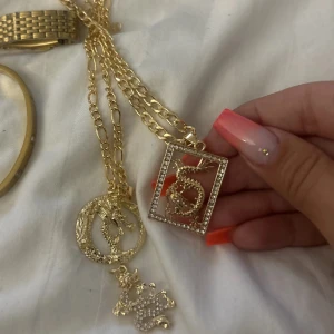 Guldiga halsband med drakmotiv - Två guldiga halsband med kedjor och hängsmycken i form av drakar och Ett hänge är inramat av en fyrkant med små glittrande stenar, det andra är runt med en drake i mitten. Perfekt för dig som gillar unika och iögonfallande smycken.