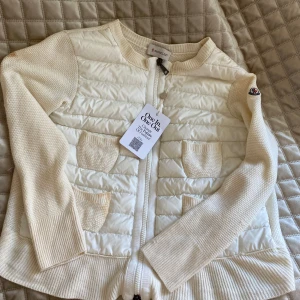 Vit quiltad cardigan jacka Moncler - Vit cardigan jacka från Moncler med quiltad front och stickade ärmar. Fyra små fickor framtill, ribbade muddar och dragkedja hela vägen. Klassisk Moncler-logga på ärmen. Ok skick, se bilder. Några fläckar men ändå fin och snygg till allt. Självklart äkta och köpt på Vestiaire. 