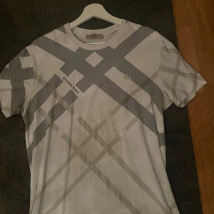 Vit rutig t-shirt från Burberry - Snygg vit t-shirt från Burberry med klassiskt rutmönster i grått och svart. T-shirten har rund hals, korta ärmar och en clean look med diskret Burberry-logga på bröstet. Perfekt för dig som gillar stilrena designer och vill sticka ut med ett ikoniskt märke.