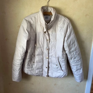 Beige quiltad pufferjacka Vero Moda - Snygg beige pufferjacka från Vero Moda med quiltad design och guldiga knappar. Jackan har hög krage, dragkedja och tryckknappar framtill samt praktiska fickor. Perfekt för kyliga dagar och enkel att matcha med olika outfits. Är lite skavd på guld färgen men inget trasigt, xl men är en L skulle ja säga 