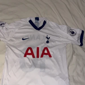 Tottenham Kane 10 matchställ Nike - Tottenham Hotspur matchställ med Harry Kane 10. Tröjan är vit med blå detaljer, kortärmad och har klubbmärke, Nike-logga och Premier League-tryck. Byxorna är marinblå med nummer 10 och Nike-logga. Materialet är lätt och andas, perfekt för fotboll.