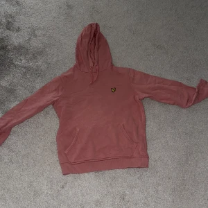 Rosa hoodie från Lyle & Scott - Säljer en snygg rosa hoodie från Lyle & Scott i storlek S. Tröjan har huva med snörning, magficka och den klassiska gula loggan på bröstet. Perfekt för chill dagar eller när du vill ha en avslappnad stil.