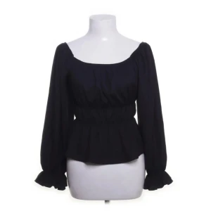 Svart blus från Holly & Whyte - Supersnygg svart blus med långa puffiga ärmar och volangdetaljer vid ärmsluten. Blusen har en smockad midja som ger en markerad siluett och en bred, rundad urringning. Perfekt för dig som gillar romantiska vibes och vill ha något stilrent men ändå trendigt.