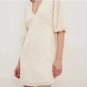 Vit kort klänning med struktur - Superfin vit kortklänning med v-ringning och puffiga trekvartsärmar. Klänningen har ett unikt strukturerat tyg med små 3D-detaljer som ger en cool look. Perfekt för dig som gillar trendiga och feminina plagg med det lilla extra.