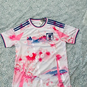 Japan fotbollströja Adidas rosa/vit - Japan landslagets fotbollströja från Adidas med unikt rosa och blått mönster inspirerat av körsbärsblommor och berg. Tröjan är vit i grunden, har korta ärmar och klassiska Adidas-ränder på axlarna. Tillverkad i lätt och ventilerande material (AEROREADY).