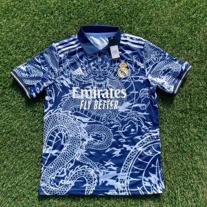 Real Madrid x Adidas blå draktröja L - Snygg Real Madrid fotbollströja från Adidas i marinblått med vitt drakmönster över hela tröjan. Klassisk krage, korta ärmar och broderade loggor. Materialet är lätt och andas, perfekt för match eller träning. Stora vita tryck på bröstet och gulddetaljer vid ärmslut.