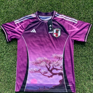Japan Adidas fotbollströja lila L - Japan fotbollströja från Adidas i lila och rosa toner med snyggt körsbärsblom-motiv och solnedgång på framsidan. Klassiska vita Adidas-ränder på axlarna, broderad logga och JFA-emblem. Slim fit, tillverkad i lätt och ventilerande polyester.