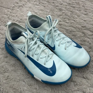 Nike Mercurial blå fotbollsskor - Nike Mercurial fotbollsskor i ljusblått och blått med vita detaljer. Skorna har snörning, låg profil och är tillverkade i syntetmaterial för bästa känsla på planen. Snygg design med stora Nike-loggor och grafiska linjer baktill. Perfekta för dig som vill sticka ut på fotbollsplanen.